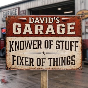 Pode incluir: Uma placa retangular de metal com acabamento desgastado apresentando o texto DAVID'S GARAGE em branco sobre um fundo vermelho escuro, seguido pelo texto KNOWER OF STUFF e FIXER OF THINGS em marrom escuro sobre um fundo creme com uma estrela.