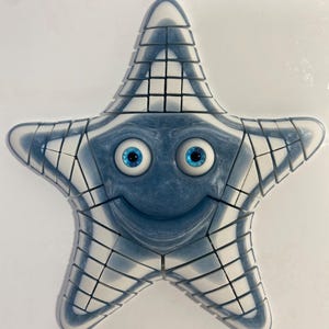 Puede incluir: Un rompecabezas azul y blanco con forma de estrella de mar sonriente con grandes ojos azules. El rompecabezas está hecho de pequeñas piezas cuadradas que encajan. La estrella de mar tiene una expresión feliz.