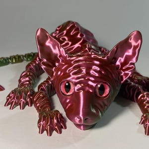 May include: A metallic red Sphynx cat sculpture with iridescent highlights. The cat is posed lying down, with large eyes and a long tail. The sculpture has a smooth, glossy finish and is a unique decorative item.