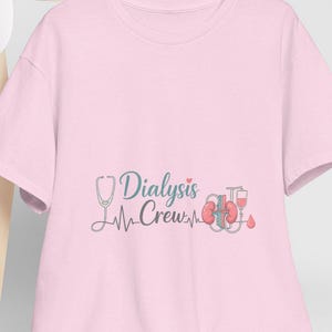 Può includere: Una maglietta rosa chiaro con la scritta "Dialysis Crew" in corsivo, insieme a uno stetoscopio, una linea del battito cardiaco e un grafico del rene. Un berretto da baseball bianco è appoggiato su una sedia di legno.