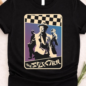 May include: Black t-shirt featuring a graphic of a band performing, with the text "The Selecter" in a stylized font. The graphic has a checkered pattern at the top and a gradient background. The shirt is made of a soft material.