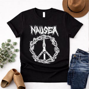 Puede incluir: Camiseta negra con la palabra "NAUSEA" en blanco, sobre un símbolo de la paz blanco rodeado por una corona de espinas. La camiseta es de cuello redondo.
