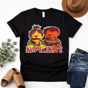 May include: Black t-shirt featuring a graphic of two puppet-like characters with orange and yellow skin, wearing striped and patterned shirts. The text "RAWPELKISTE" is printed in red and white below the characters.