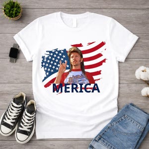 May include: White t-shirt featuring a graphic of a man with a mullet and the word "MERICA" in navy blue. The design includes a stylized American flag in red, white, and blue. The man is making a rock and roll hand gesture.