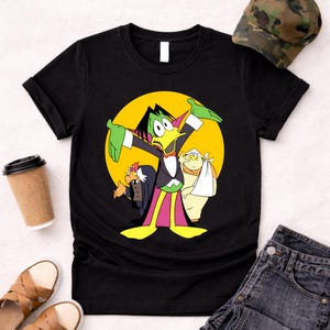 Op de afbeelding: Zwart T-shirt met een stripfiguur in smoking en een gele achtergrond. De afbeelding bevat twee andere stripfiguren. Een camouflage hoed, spijkerbroek shorts en sandalen zijn ook zichtbaar.