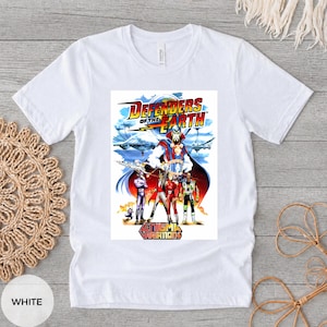 May include: White t-shirt with a vibrant graphic from the cartoon "Defenders of the Earth." The design includes the show's title, characters, and the words "Enigma Variations."