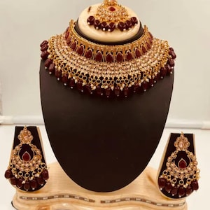 Peut inclure: Un ensemble de bijoux doré comprenant un collier, des boucles d'oreilles et un serre-tête. Le collier et les boucles d'oreilles présentent un design de perles et de cristaux marron. Le serre-tête est rond avec une pierre marron.