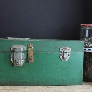 May include: A rectangular green metal toolbox with three silver-colored metal latches on the front. Next to the toolbox are three clear glass jars with red and white lids, each filled with various metal hardware components and small fasteners.