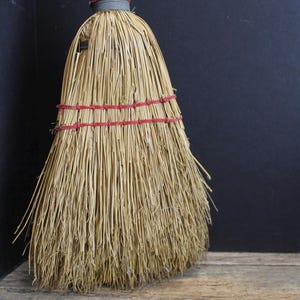 May include: A straw broom with a red handle and gray binding. The broom's straw bristles are bound with red twine. The handle is red with some paint chipping. The broom is set against a dark background.