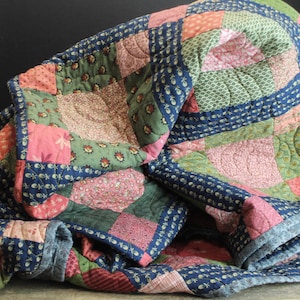May include: A patchwork quilt featuring a variety of square fabric blocks in shades of navy blue, sage green, and dusty pink. The quilt displays intricate floral and geometric patterns with visible machine stitching throughout the textile surface.