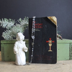May include: A white porcelain angel figurine stands on a green wooden box. A black book with the title "Sunday Missal" is on the box. A silver rosary with a silver crucifix hangs from the book.