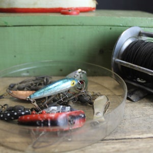 May include: A vintage fishing reel with black line, a green wooden box, and a clear plastic dish filled with fishing lures, including a blue and silver lure, a copper lure, and red and black lures.