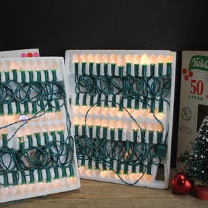 Pode incluir: Conjuntos de luzes de Natal vintage com fios verdes e lâmpadas brancas, exibidos em sua embalagem original. A imagem também inclui uma pequena árvore de Natal, enfeites e uma caixa que diz "Yuletide".