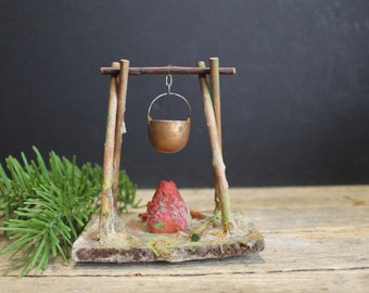 May include: A miniature campsite with a small copper pot hanging over a red fire pit. The campsite is made of wood and has a rustic look.