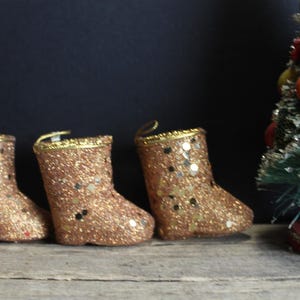 May include: Three glittery gold Christmas stocking ornaments with gold trim and a small Christmas tree with red and green decorations. The ornaments are on a wooden surface.