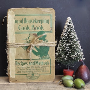 May include: A vintage cookbook titled "Good Housekeeping Cook Book" with a green illustration of a woman on the cover. The book is tied with twine and has the text "Recipes and Methods for Every Day and Every Occasion" on the bottom.