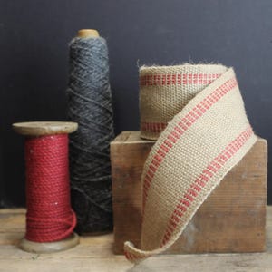 May include: A still life arrangement featuring yarn spools and a roll of burlap ribbon. The yarn spools are red and grey. The burlap ribbon is beige with red stripes. The items are on a wooden surface.