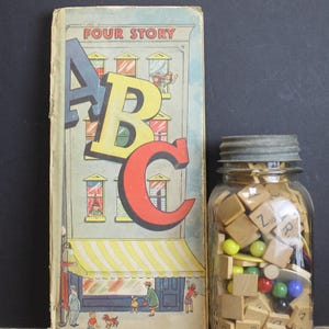 May include: Vintage children's book titled "Four Story ABC" with a colorful illustration of a building and large letters. Beside it is a glass jar filled with wooden letter tiles and colorful marbles.