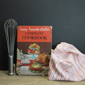 May include: A red and white striped kitchen towel sits next to a vintage cookbook titled "Amy Vanderbilt's Complete Cookbook". A silver whisk sits in front of the cookbook.