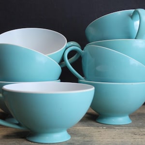 May include: Six turquoise ceramic teacups with white interiors are stacked. Each cup has a curved handle and a rounded base. The cups are arranged on a wooden surface against a dark background. The cups are a solid colour.