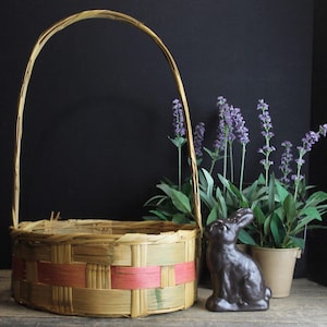 May include: A round woven basket with a pink and brown trim and a handle. The basket is empty and sitting on a wooden surface. There is a small brown rabbit figurine and a potted plant with purple flowers in the background.