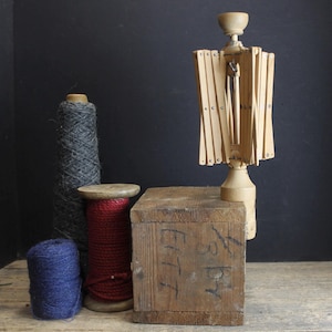 May include: A still life composition featuring a wooden yarn swift, spools of yarn in blue, red, and gray, and a wooden box with faded markings. The yarn swift is light-colored and stands upright, while the yarn spools are various sizes.