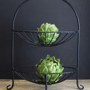 May include: A two-tiered black metal fruit basket with a handle, holding two green artichokes. The basket has a curved design and is placed on a wooden surface against a dark background. The artichokes are fresh and vibrant.