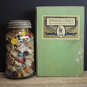 Può includere: Un barattolo di vetro pieno di tessere di lettere di legno colorate e un libro verde intitolato "Pinocchio: Le avventure di un burattino".