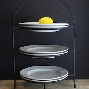 May include: A three-tiered black metal stand featuring a curved handle at the top. Each of the three levels holds a stack of two white ceramic plates. A single bright yellow lemon is placed centrally on the top plate of the highest tier.
