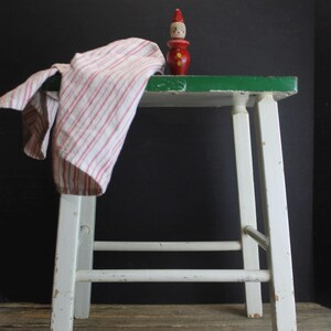 May include: A white and green painted wooden stool with a red and white striped cloth draped over the side. A small, red wooden clown figurine with a white face and a pointed hat sits on top.