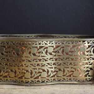 May include: An antique brass-toned rectangular container with intricate floral cutouts. The container has a copper-colored interior and sits on a wooden surface. A shiny red Christmas ornament is next to the container.
