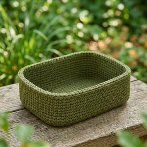 Könnte beinhalten: Ein rechteckiger, olivgrüner Häkelkorb steht auf einer verwitterten Holzoberfläche. Die Seiten des Korbs sind dick und strukturiert, mit einem sichtbaren Stichmuster. Der Hintergrund zeigt verschwommenes grünes Laub.