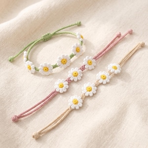 May include: Three daisy chain bracelets in the image. One is green, and the others are pink and tan. Each bracelet features white daisy flowers with yellow centers. The bracelets are adjustable and made of a soft, woven material.