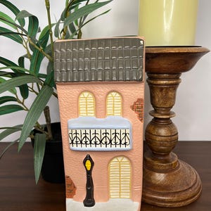 May include: A ceramic house-shaped candle holder in shades of pink, gray, and white. The facade features arched windows, a balcony with a black railing, and a black lamppost. A wooden candle holder and a green plant are in the background.