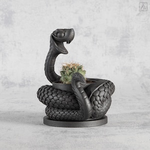 Peut inclure: Un pot de plante noir en forme de serpent avec un cactus à l'intérieur. Le pot a une texture, un corps enroulé, une tête relevée et une base ronde. Le cactus est vert avec des épines.