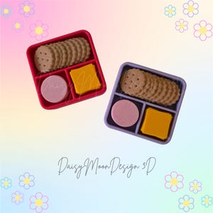 May include: Two square divided snack containers, one red and one purple, each holding a stack of round tan crackers, a single pink circular disc, and a stack of yellow square slices. The text Daisy Moon Design 3D is visible in the center.
