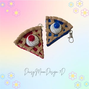 May include: Two pie slice keychains. One is red with a white dollop and a red cherry on top. The other is blue with a white dollop and a blue blueberry on top. Both have a tan crust and a silver clasp.