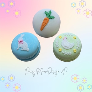 May include: Three decorative macarons with Easter-themed designs. One is light blue with a white bunny, another is white with a carrot, and the last is light green with a white chicken. The background is pastel with floral accents.