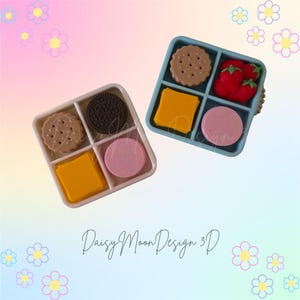 May include: Two square, four-compartment snack trays. The left tray is light pink and contains a tan cookie, a dark brown sandwich cookie, a yellow square, and a pink circle. The right tray is blue and holds a tan cookie, red strawberries, a yellow square, and a pink circle.