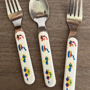 May include: Two stainless steel forks and one stainless steel spoon with white handles. Each handle features a repeating pattern of colorful small vehicles including a red train, a blue boat, a blue airplane, and a yellow car.