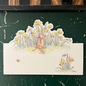 May include: A whimsical greeting card featuring a watercolor illustration of a teddy bear seated among yellow daffodils. The card has a scalloped top edge and a blank space for a message. The bear has a red bow.