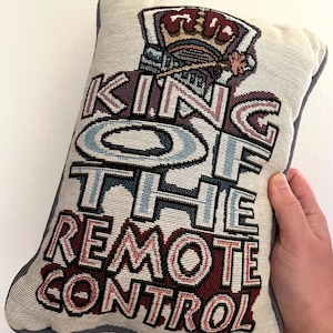 May include: Rectangular decorative pillow with the text "KING OF THE REMOTE CONTROL" in a tapestry style. The text is in shades of red, blue, and purple, with a crown graphic at the top. The pillow has a light beige background and a gray border.