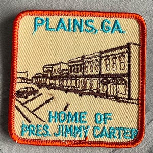 May include: Embroidered patch with the text "PLAINS, GA. HOME OF PRES. JIMMY CARTER" in turquoise. The patch features a brown illustration of a street scene and is bordered with orange stitching.