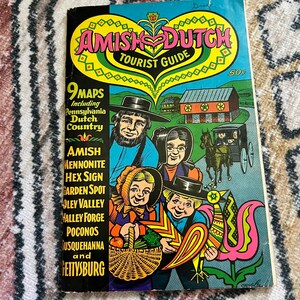 May include: A vintage "Amish Dutch Tourist Guide" book cover. The cover features a colorful illustration of an Amish family, a horse-drawn carriage, and a farm scene. The title is in large, stylized letters, with the price of 50 cents in the upper right corner.
