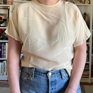 May include: A cream-colored, short-sleeved blouse with a round neckline. The blouse is made of a lightweight, slightly sheer fabric. The model is wearing blue denim jeans.