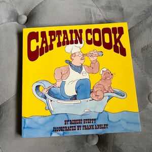May include: A children's book titled "Captain Cook" with a yellow cover. The illustration depicts a cartoon chef with a spyglass in a teacup boat, accompanied by a cat. The book's title is in large, maroon letters.