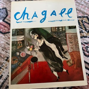 May include: A book cover with the title "Chagall" in blue cursive lettering. The cover features a painting of a couple in a room with a red floor, a window, and a bouquet of flowers.
