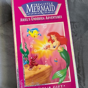 May include: A pink VHS box for Disney's The Little Mermaid: Ariel's Undersea Adventures. The box features Ariel, Flounder, and Sebastian. The title is in a blue and white font. The box reads "Ariel's Gift."