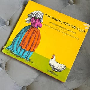 May include: A vintage children's book titled "The Woman with the Eggs" by Hans Christian Andersen. The cover features a colorful illustration of a woman in a long dress and a white chicken on a green field, set against a bright yellow background.