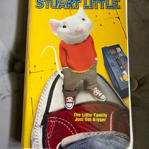 May include: A Stuart Little VHS tape featuring a white mouse character wearing a red sweater and tan pants, standing on a large red and white sneaker. The yellow cover includes the text Stuart Little, From the co-director of The Lion King, and The Little Family Just Got Bigger, along with a blue suitcase.